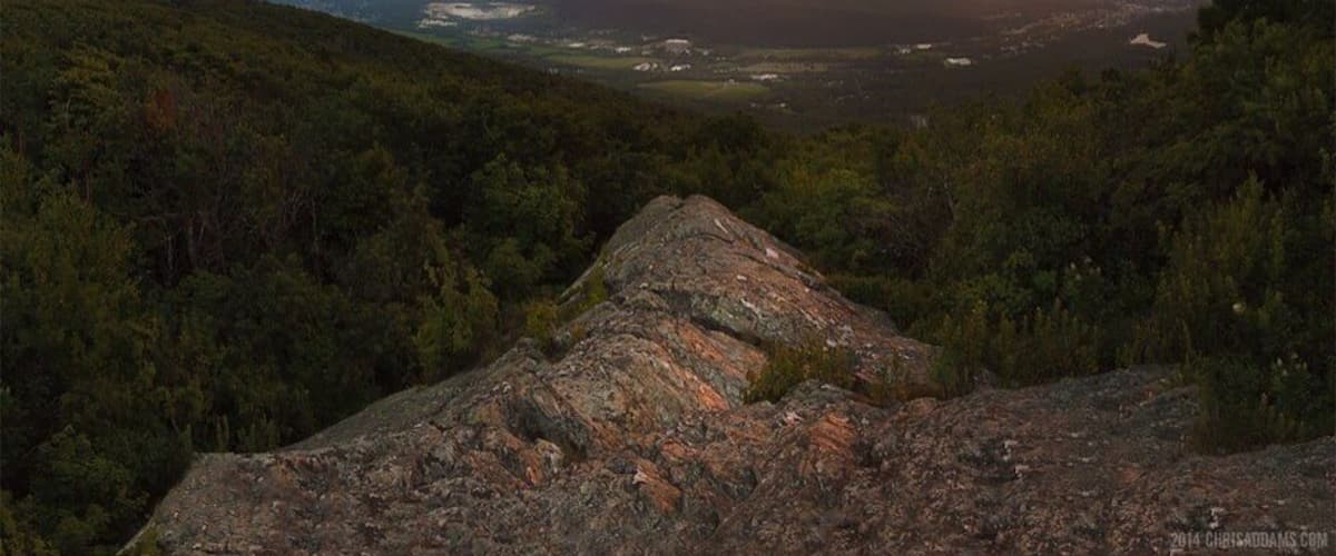 A mountainous landscape with a prominent rock formation in the foreground and a valley with scattered buildings in the distance under a sunset sky.