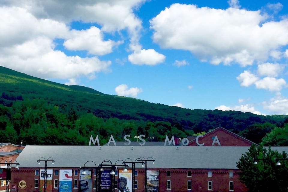 En byggnad med texten ”MASS MoCA” på fasaden, omgiven av grönska och kullar.