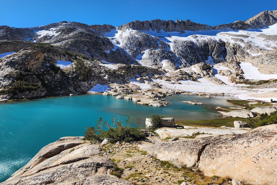 Un paisaje montañoso con un lago turquesa rodeado de terreno rocoso y parches de nieve.