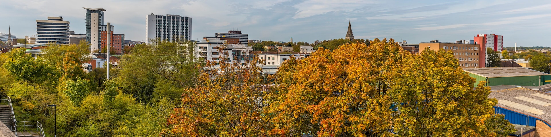 Une vue urbaine avec des arbres aux couleurs automnales, des bâtiments modernes et le clocher d’une église.