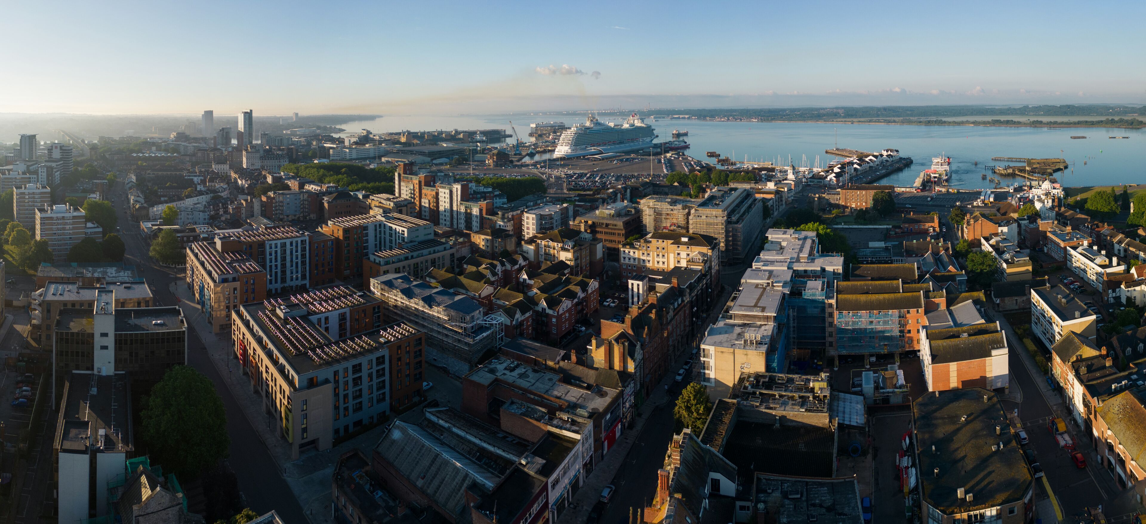 Southampton UK seaside city panorama. Wide angle aerial view from the ferry terminal and port with large cruise ship mooring towards the Itchen toll bridge. Beautiful sunrise and calm sea. Urban area.