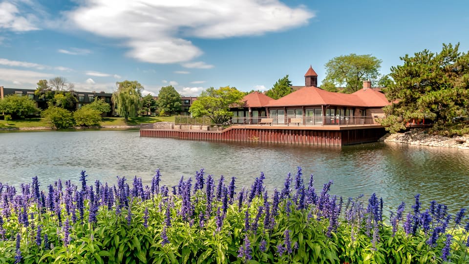 A lakeside building with a red roof, surrounded by lush greenery and purple flowers.