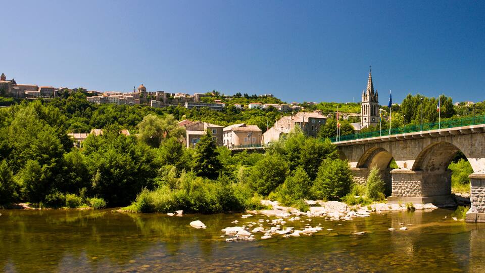 A stone bridge over a river, surrounded by greenery and buildings in the background.
