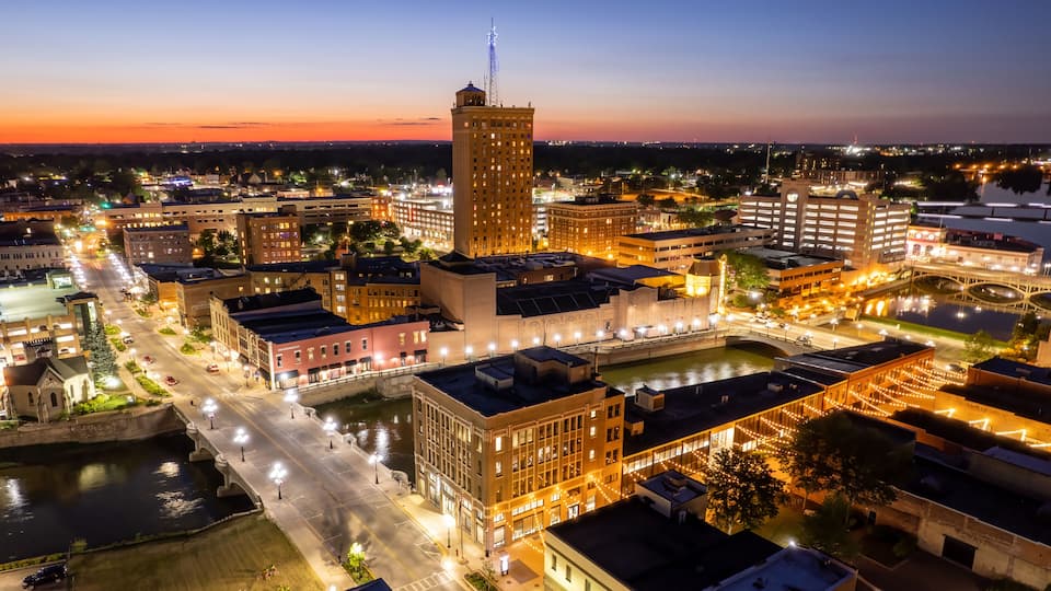 A cityscape at dusk with illuminated buildings, a prominent tower, and a river running through the center.