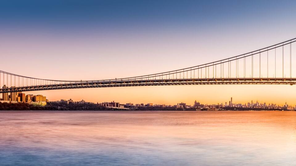 A suspension bridge spanning a river with a city skyline in the background.