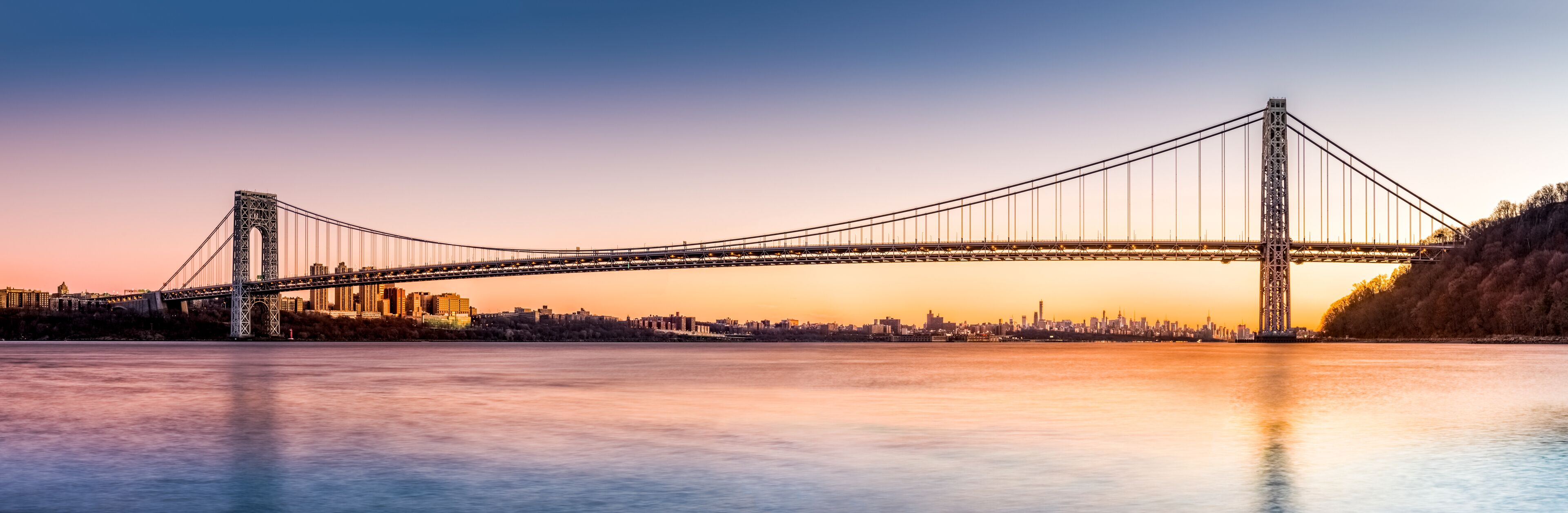 A suspension bridge spanning a river with a city skyline in the background.