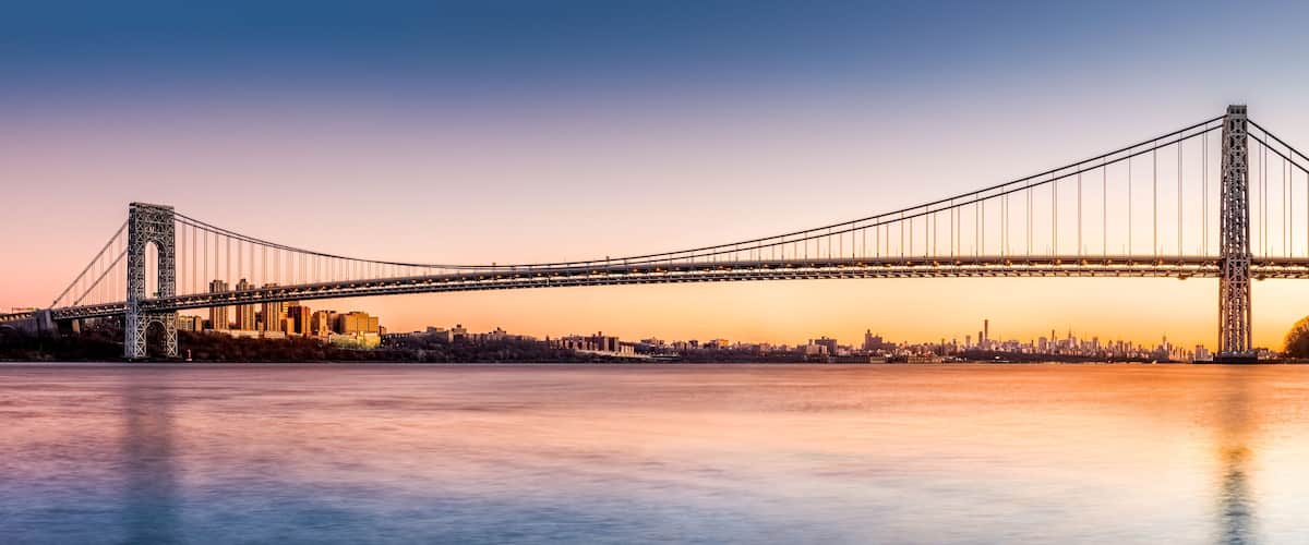 A suspension bridge spanning a river with a city skyline in the background.