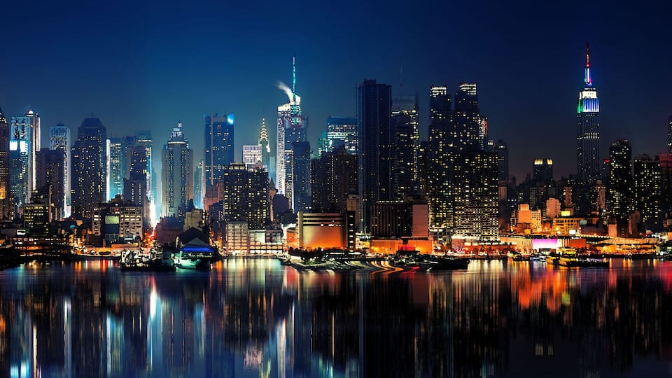 A nighttime cityscape with illuminated skyscrapers and a river reflecting the lights.