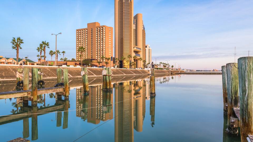 A calm waterfront with reflections of buildings and palm trees.
