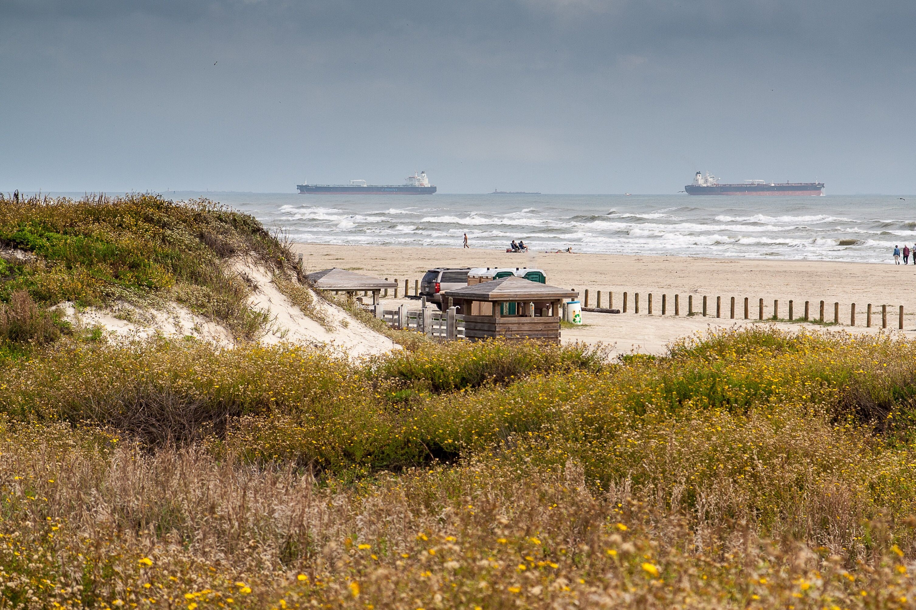 Una playa con dunas, una torre de salvavidas y dos grandes barcos a lo lejos.
