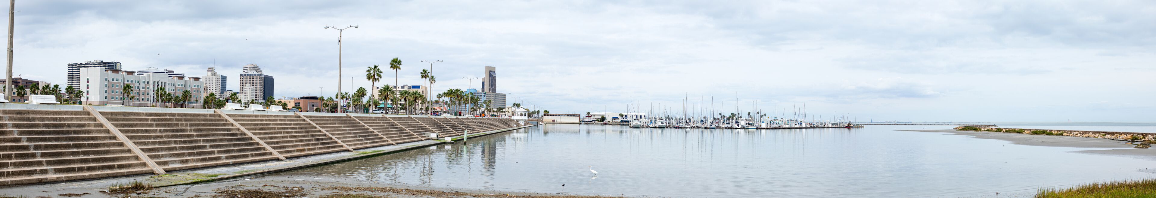 Un paseo marítimo con escalinatas que conducen a un puerto deportivo, palmeras y edificios modernos al fondo.