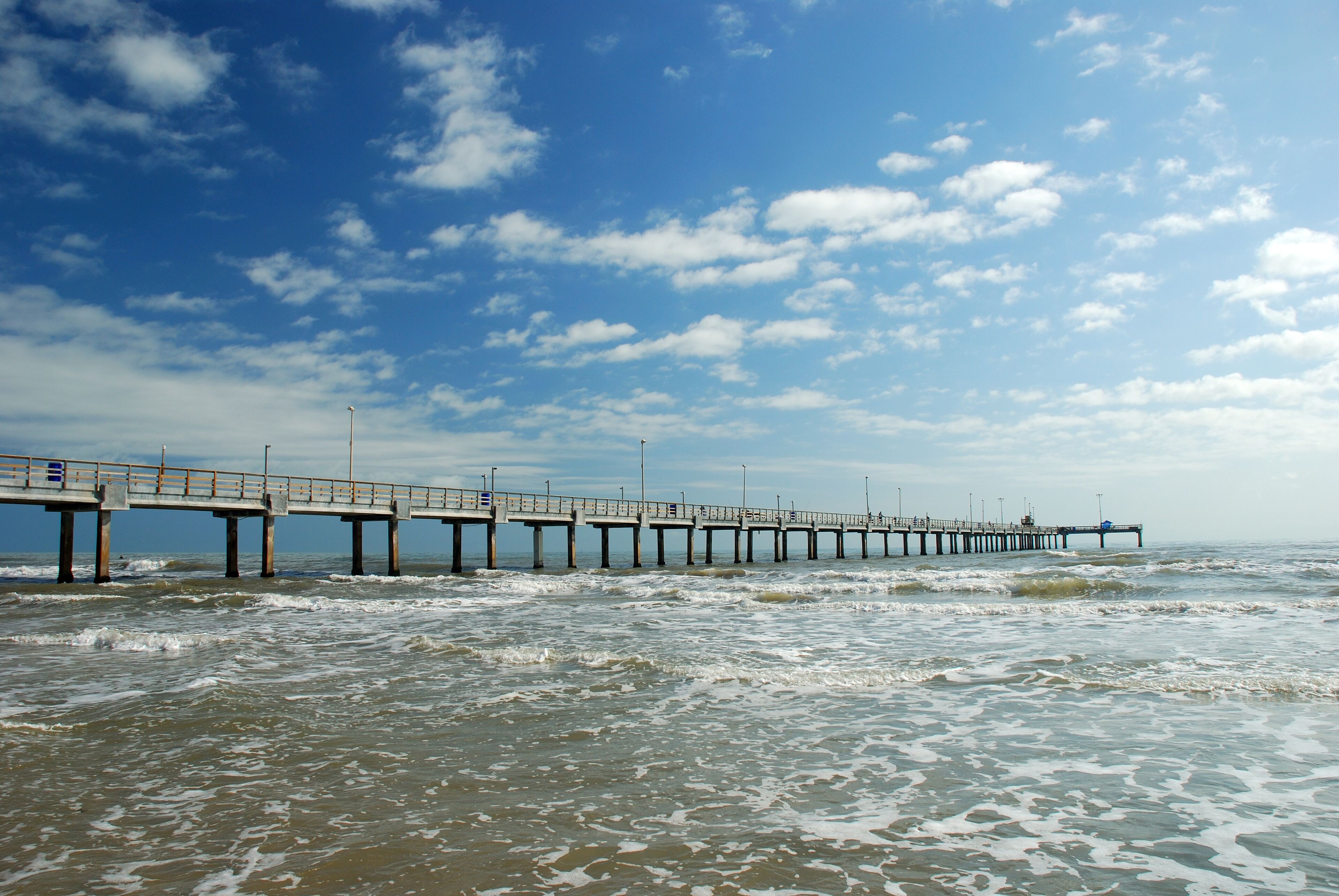 Un muelle se adentra en el océano con olas rompiendo a su alrededor bajo un cielo parcialmente nublado.