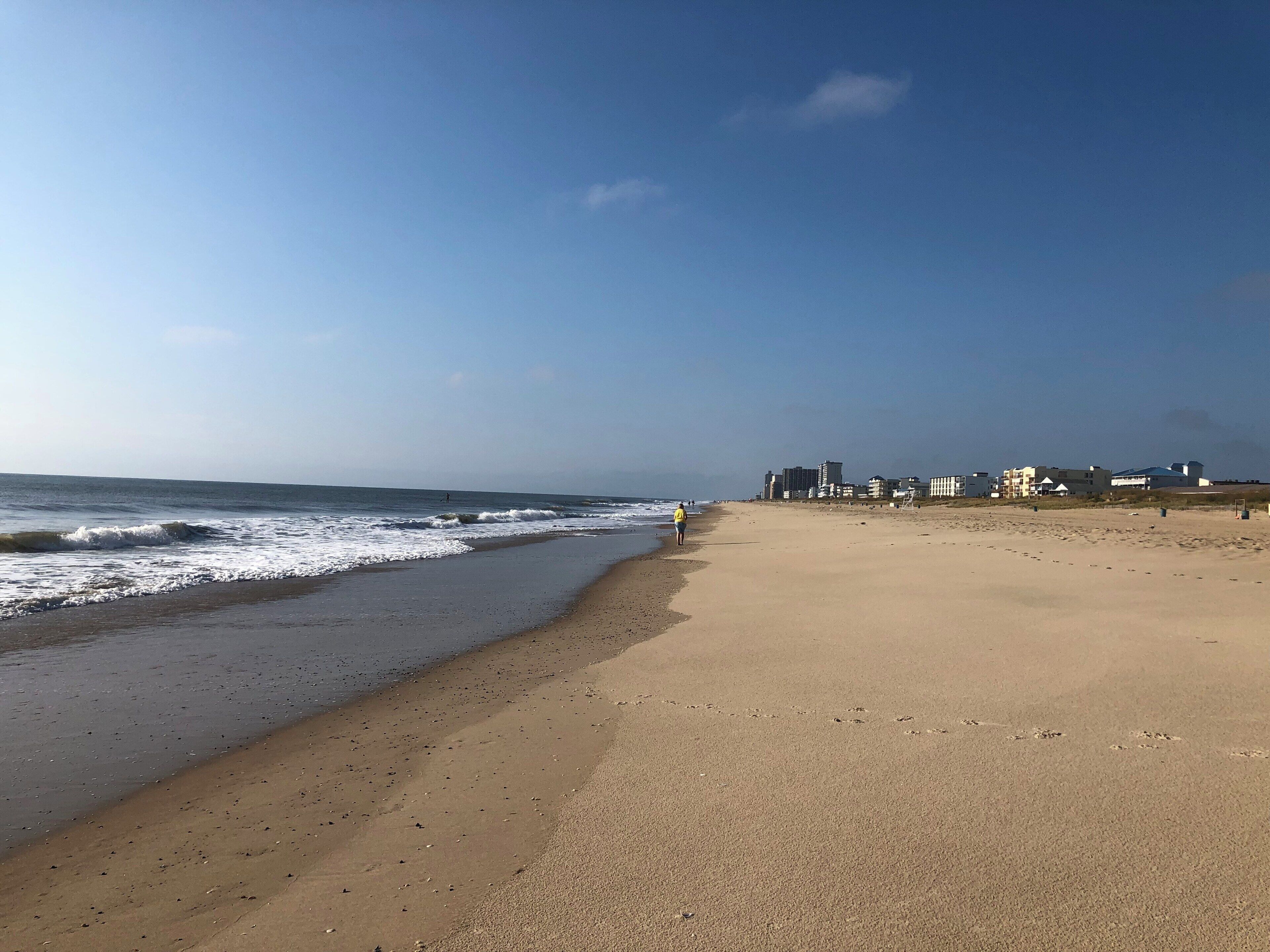 A sandy beach with waves, a clear sky, and buildings in the background.