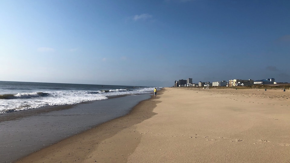 A sandy beach with waves, a clear sky, and buildings in the background.