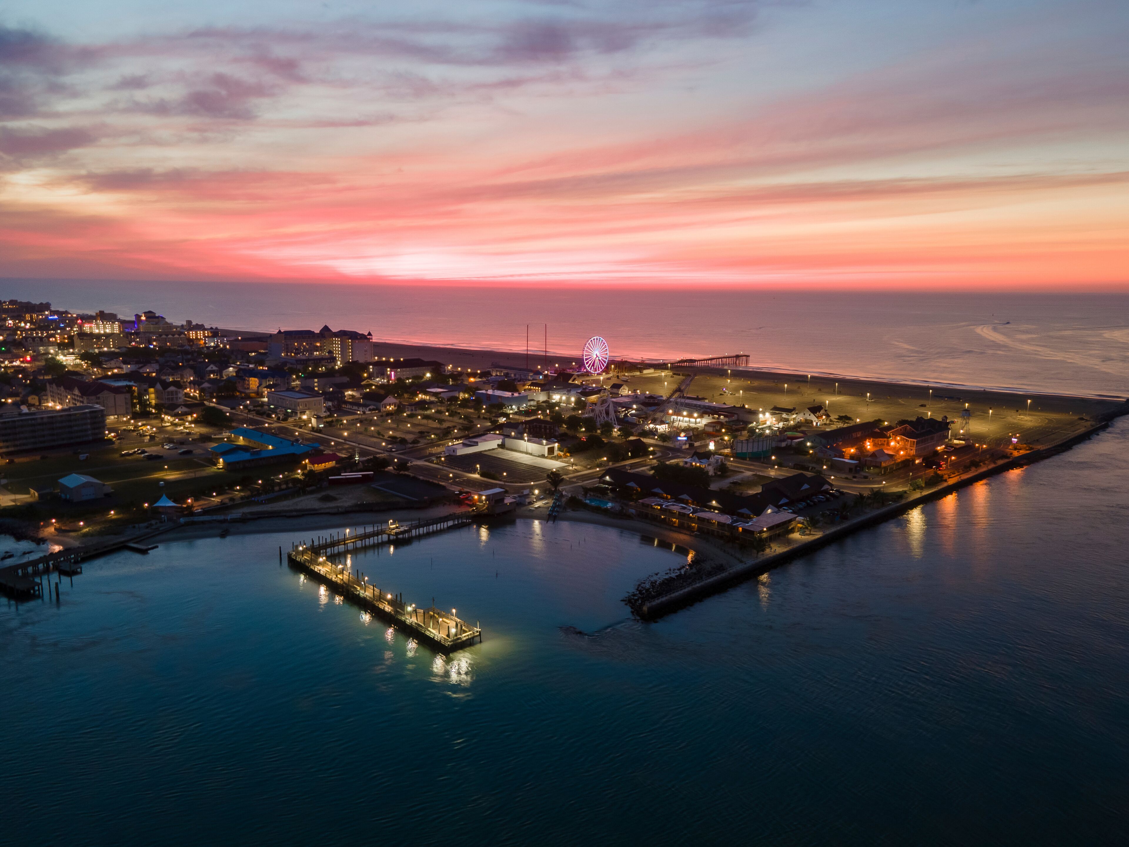 A coastal cityscape at dusk with a prominent pier and a lit Ferris wheel.