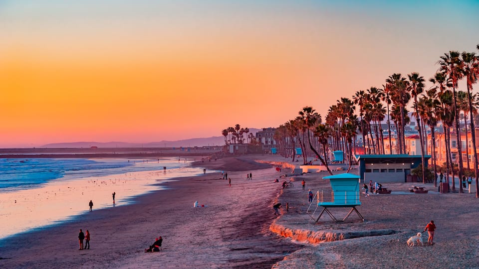 A beach at sunset with palm trees, a lifeguard tower, and people walking along the shore.