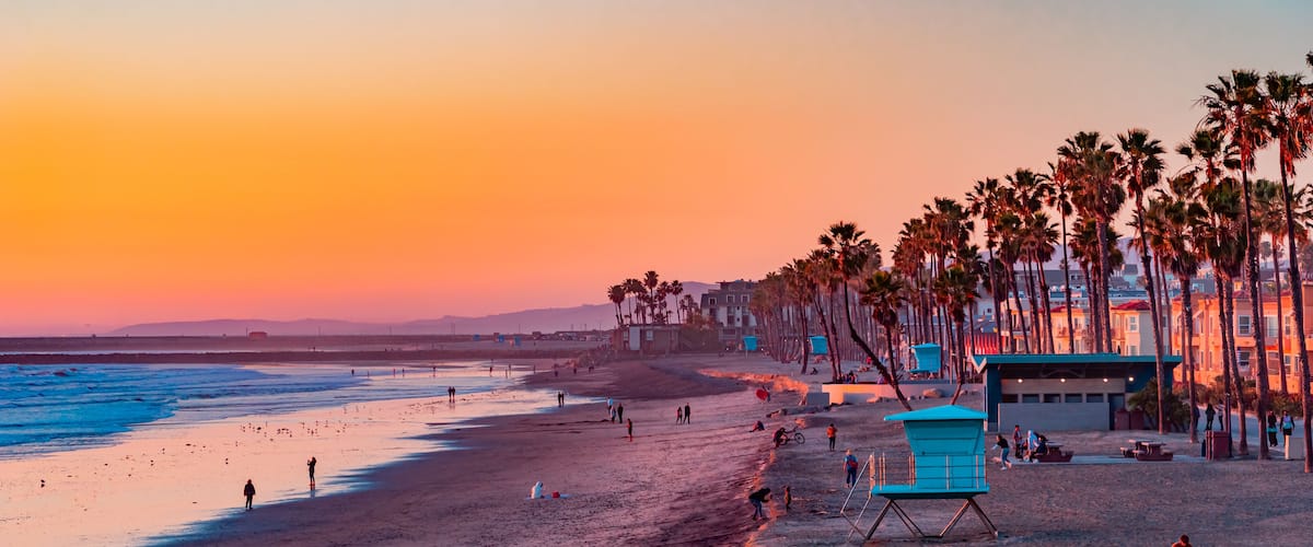 A beach at sunset with palm trees, a lifeguard tower, and people walking along the shore.