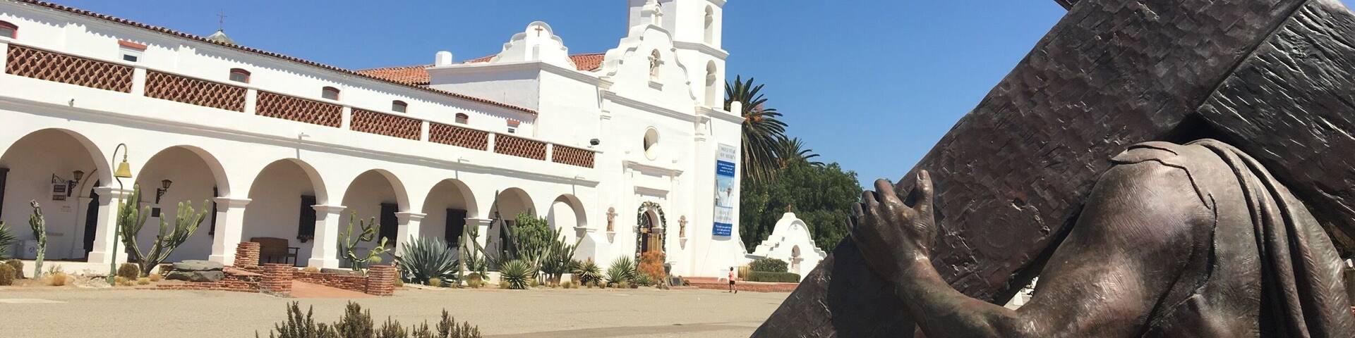 Una estatua de Jesús cargando una cruz frente a una iglesia blanca con arcos y un campanario.