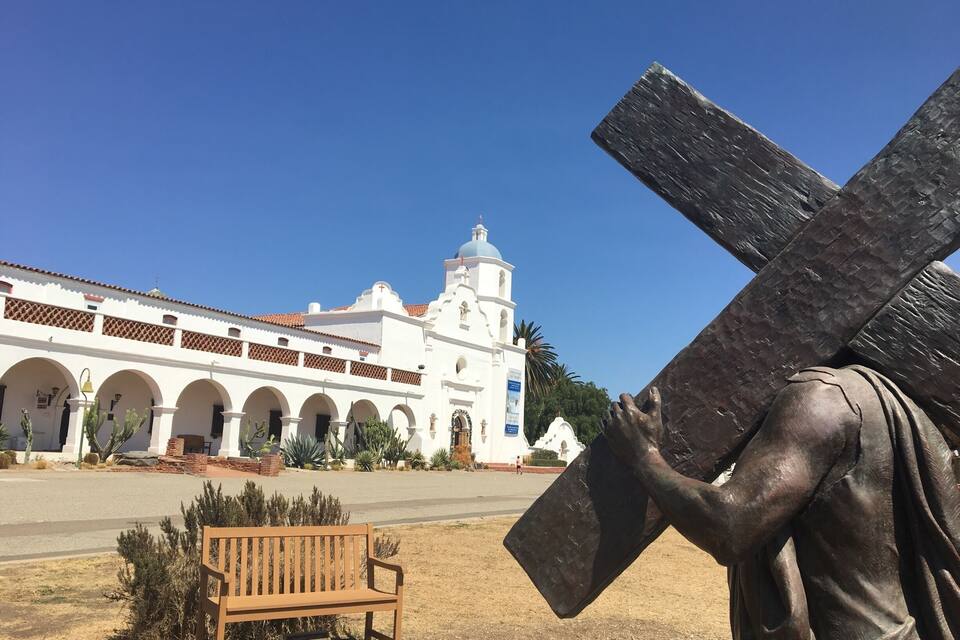 A statue of Jesus carrying a cross in front of a white church with arches and a bell tower.