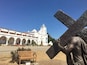 A statue of Jesus carrying a cross in front of a white church with arches and a bell tower.