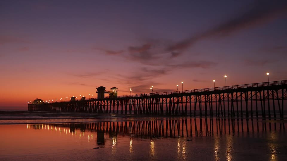 A pier extending into the water at sunset with lights along its length and a building on the left.