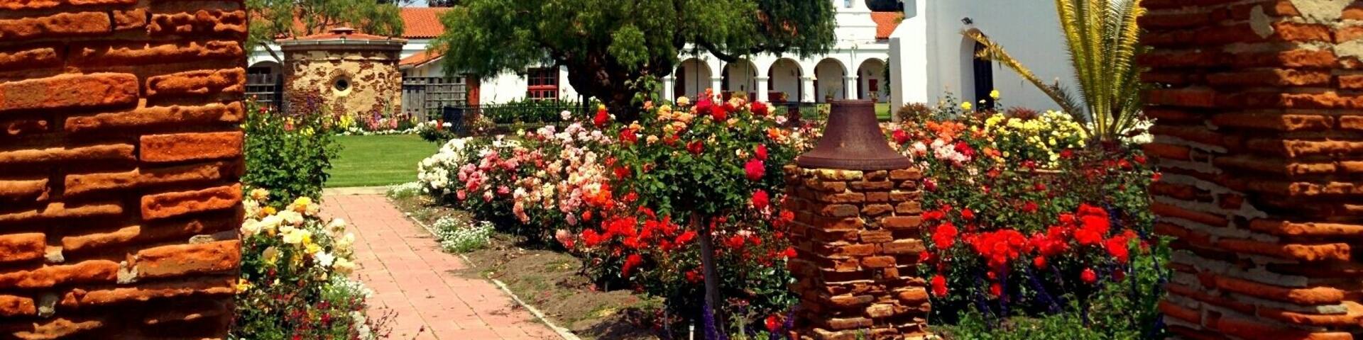 An arched brick structure framing a garden with colorful flowers and a white building in the background.