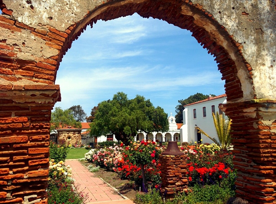 An arched brick structure framing a garden with colorful flowers and a white building in the background.