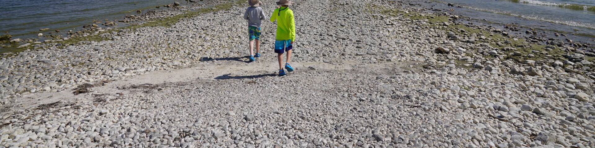 A pebbly beach with two people walking towards a forested island in the distance.