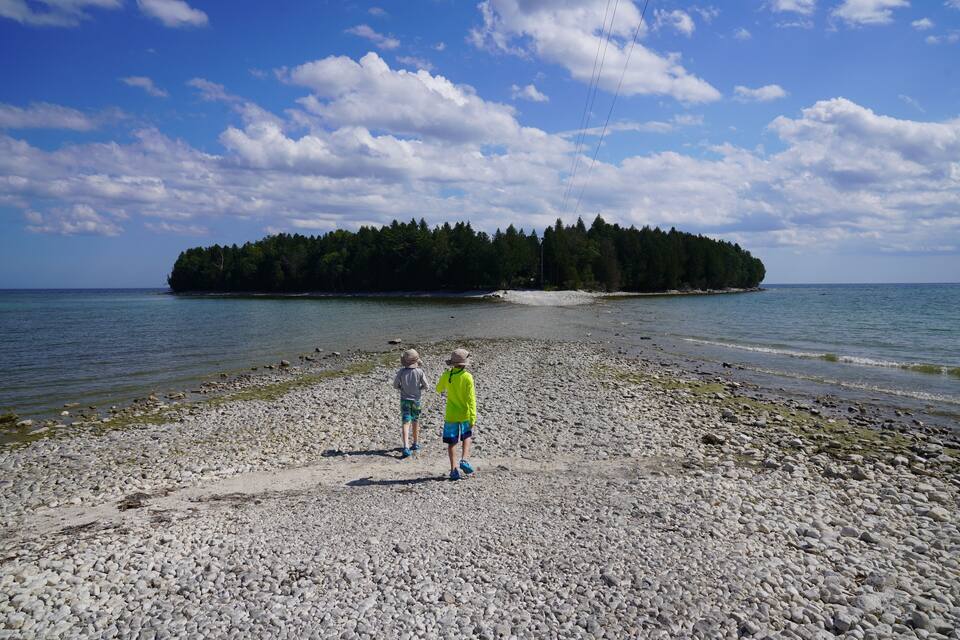 Een kiezelstrand met twee mensen die richting een bebost eiland in de verte lopen.