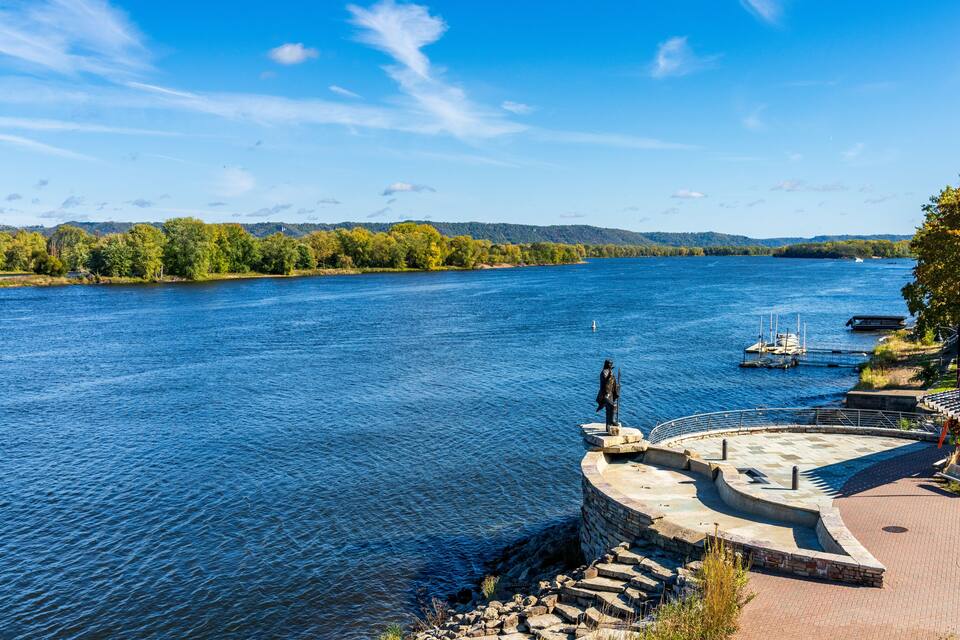 A river with a statue on a stone platform, surrounded by trees and hills.