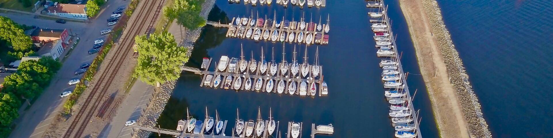 Une vue aérienne d’une marina où de nombreux bateaux sont amarrés à des quais en bois, le tout étant entouré d’une voie ferrée et d’une route bordée de voitures garées.