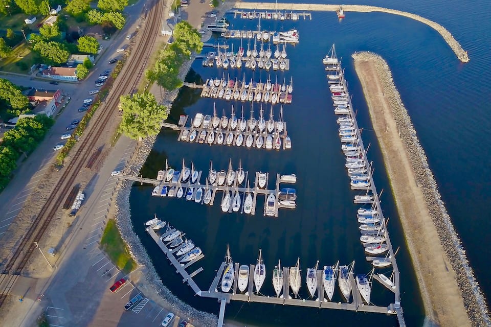 Vista aérea de un puerto deportivo con numerosos barcos amarrados a muelles de madera, rodeado por una vía de tren y una carretera con coches aparcados.