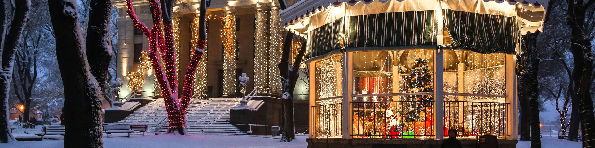 A snow-covered park with a gazebo and a building adorned with lights.