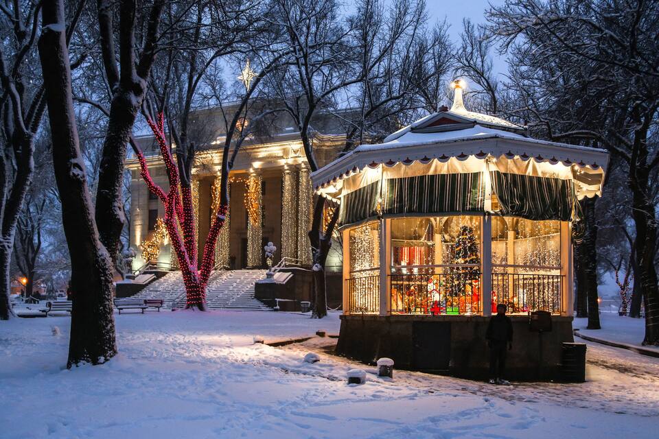 Um parque coberto de neve com um coreto e um edifício decorado com luzes.