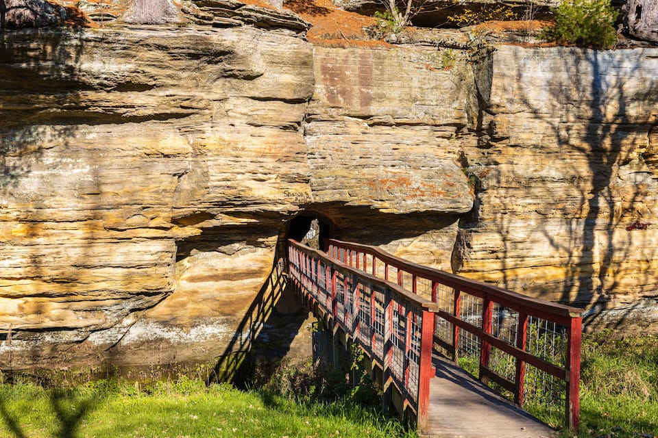 A stone bridge with a red railing leading to a cave entrance in a rocky cliff.