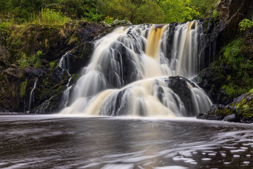 Een waterval stort over rotsen in een rivier, omringd door weelderige vegetatie.