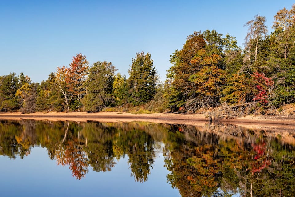 Un lac paisible où se reflètent les arbres aux couleurs automnales, entouré d’une zone boisée.