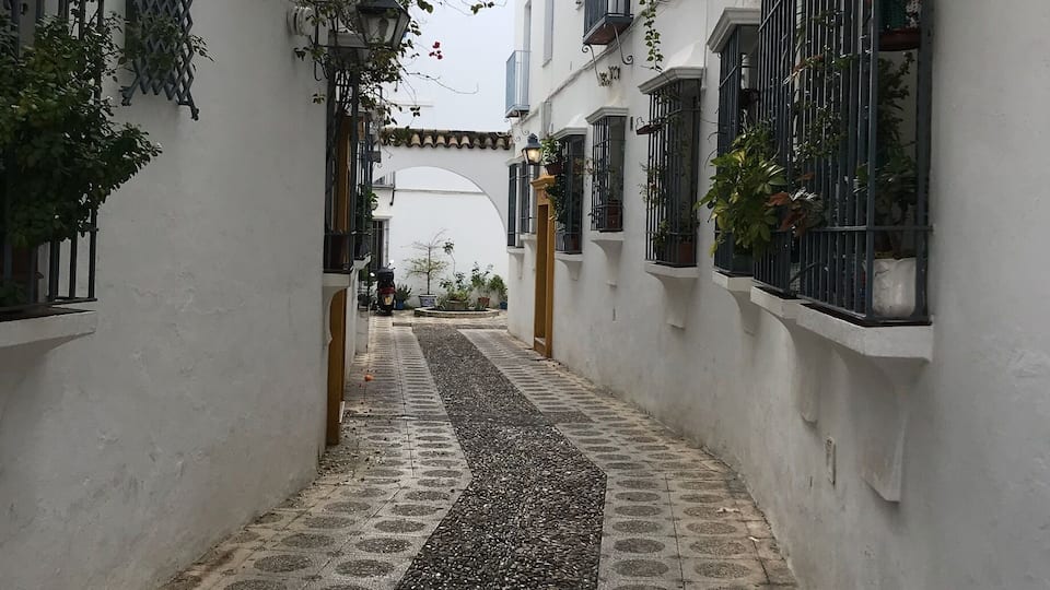 Une ruelle étroite, avec un chemin pavé, des bâtiments blancs et un pont qui se dessine au loin.