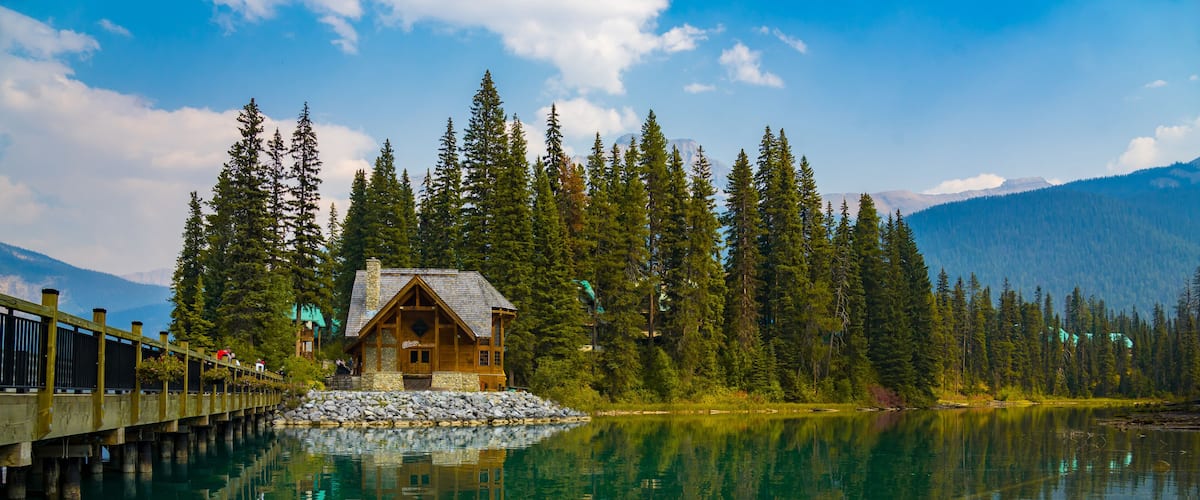 A wooden cabin by a calm lake surrounded by tall pine trees and mountains.