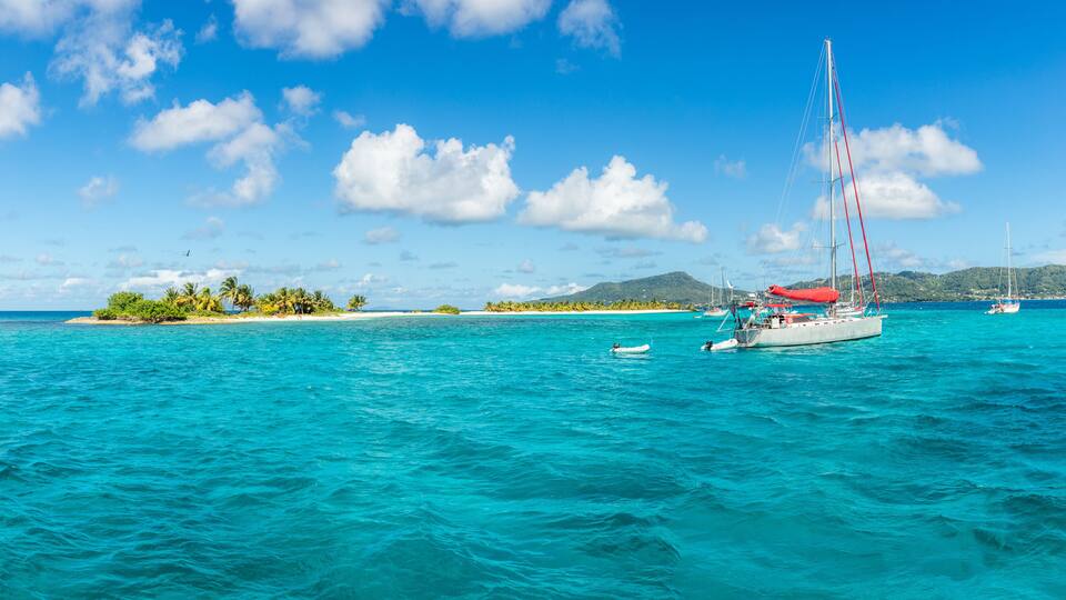 A turquoise sea with a sailboat, a small island with palm trees, and a clear blue sky.
