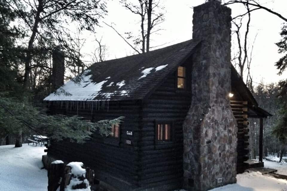 Una cabaña de troncos con chimenea de piedra, rodeada de nieve y árboles.