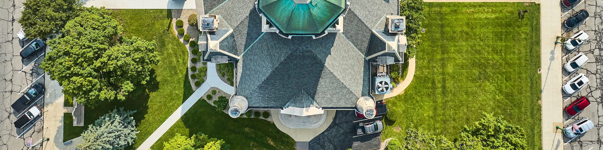 An aerial view of a park with a central gazebo, surrounding pathways, and parked cars.