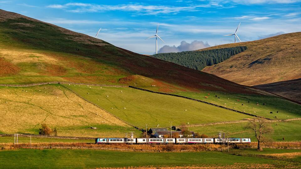 A train in a rural landscape with rolling hills, wind turbines, and grazing sheep.