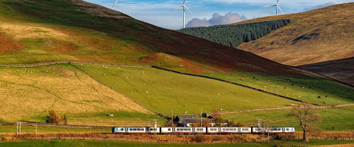 A train in a rural landscape with rolling hills, wind turbines, and grazing sheep.