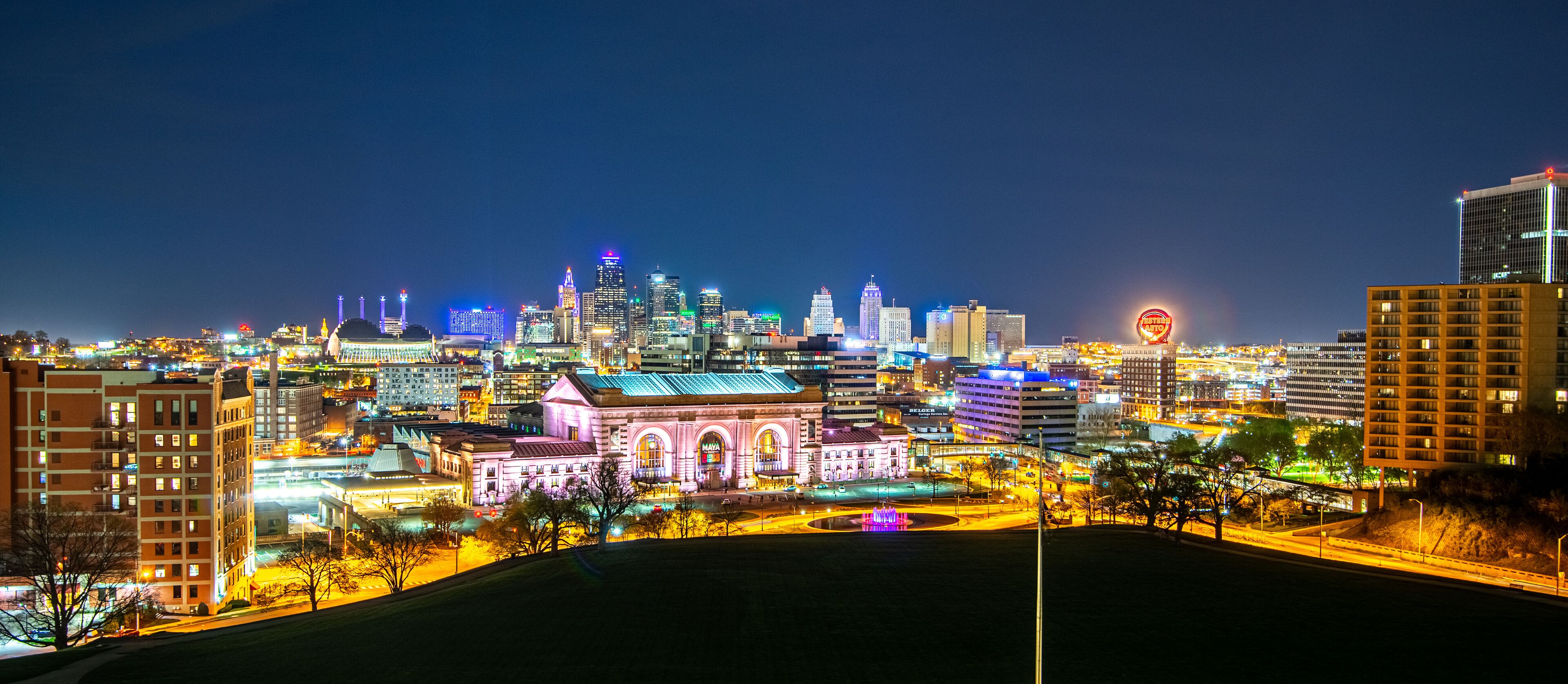 A city skyline at night with illuminated buildings and a prominent pink-lit building in the foreground.