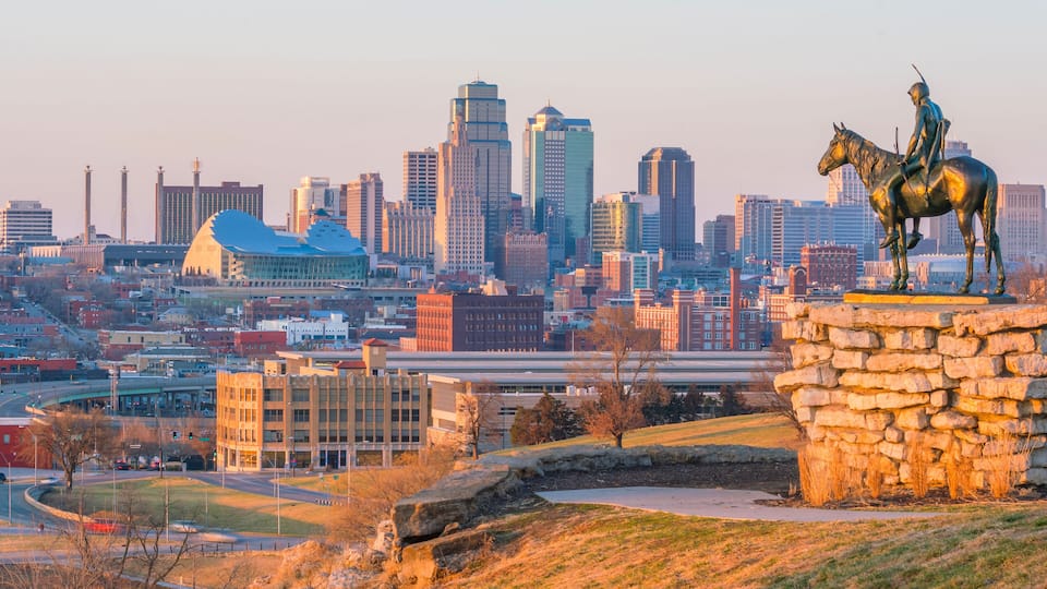 A city skyline with modern buildings, a statue of a person on a horse, and a stone wall.