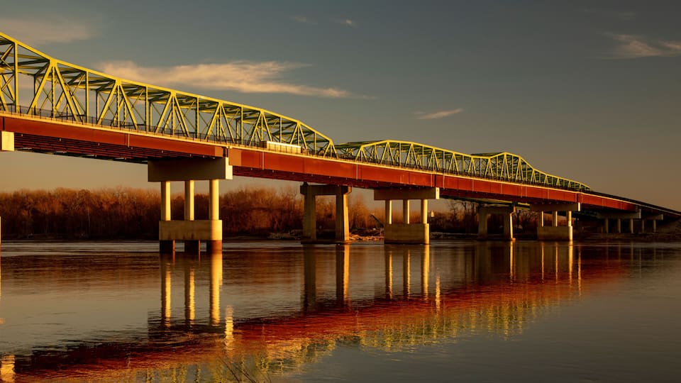 A steel bridge with yellow trusses spanning over a calm river, with bare trees in the background.