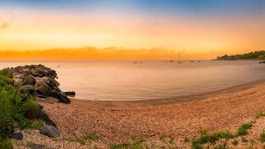 A pebbled beach with a rocky outcrop, a bridge in the background, and a sunset over the water.