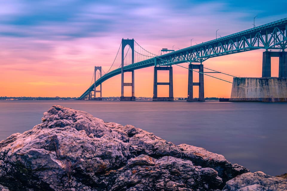 A suspension bridge over calm water at sunset with rocky foreground.