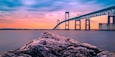 A suspension bridge over calm water at sunset with rocky foreground.